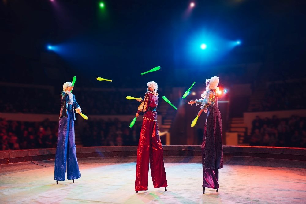 Three circus performers juggling glowing batons under colorful stage lights, showcasing a theatrical and dynamic spectacle. This image could metaphorically represent the multifaceted challenges of managing AI customer experience in a complex, fast-paced environment.