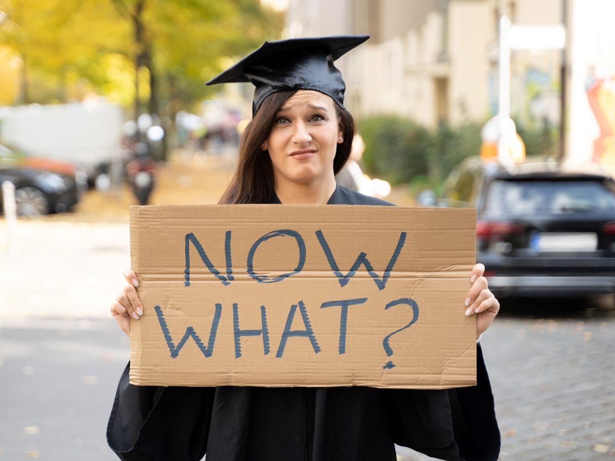 College graduate in cap and gown standing on a city street holding a cardboard sign reading “NOW WHAT?” with a frustrated, uncertain expression.