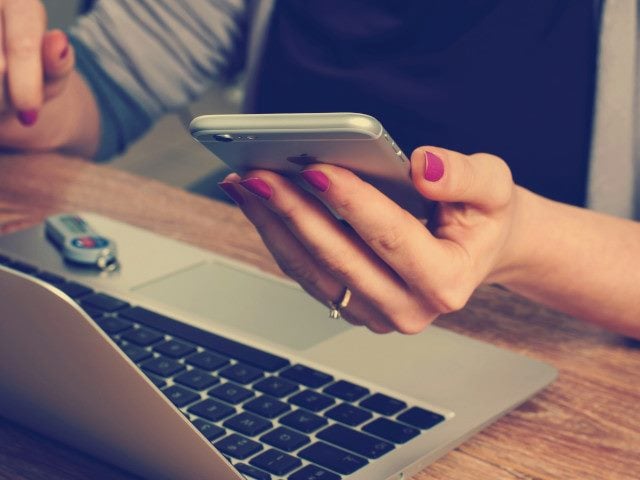 A customer holds a mobile phone in their hand at a desk with a laptop computer.