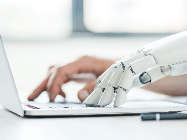 A robot hand and human hand type on a laptop simultaneously, indicating the importance of AI in customer experience and employee experience. 
