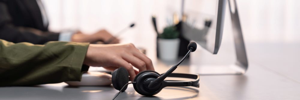Panorama focus hand holding headset on call center workspace desk with blur background of operator team or telesales representative engaging in providing client with customer support service.