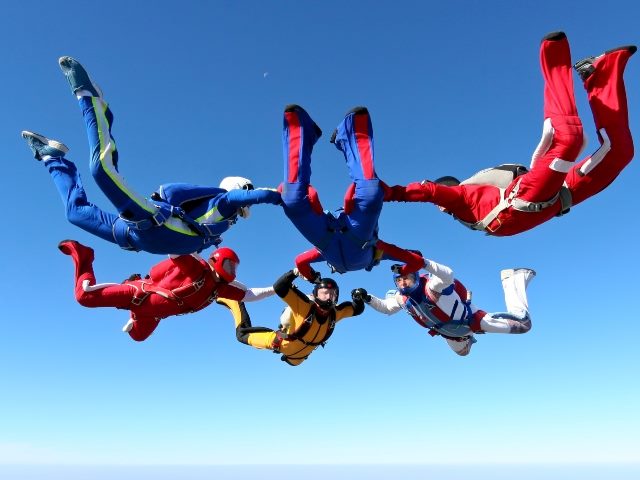 Six skydivers wearing red, white, blue and yellow skydiving suits form a circle by holding hands.