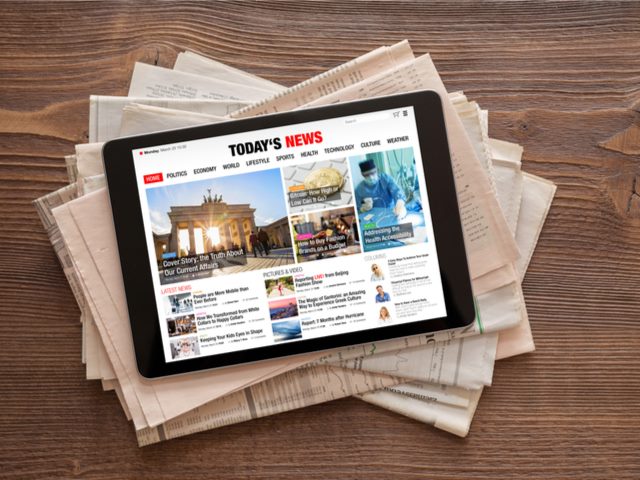 A tablet with a news app open sitting on a table atop a pile of newspapers