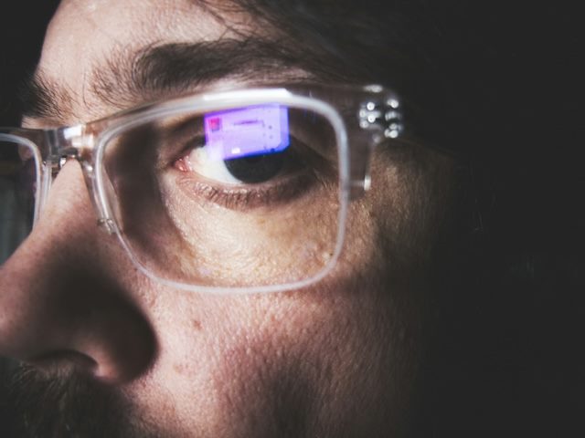 close up of a man wearing glasses, looking wary, with visible reflection of his laptop in glasses