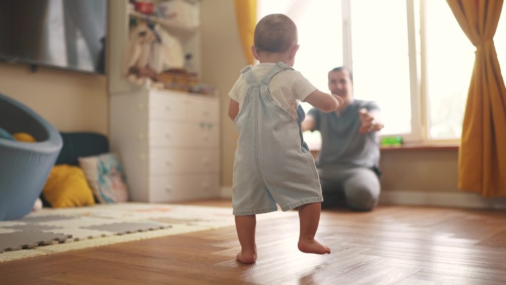 A toddler takes their first steps towards a smiling parent who is encouraging them with open arms, illustrating the concept of action-based artificial general intelligence, where learning and adaptation occur through physical interaction and movement.