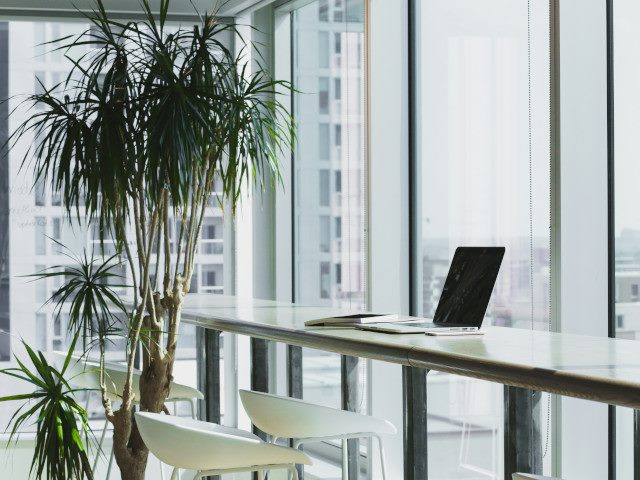 A laptop computer on an office table beside a high-rise window.