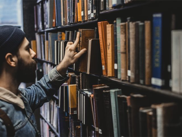 A man wearing a denim jacket and beanie reaches his hand out to examine a book on a full library shelf