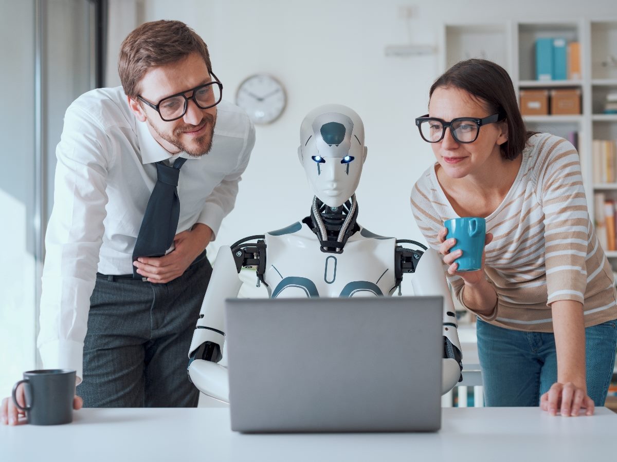 A humanoid robot sits at a desk working on a laptop while two human coworkers lean in beside it, smiling and observing its screen during a collaborative office session.