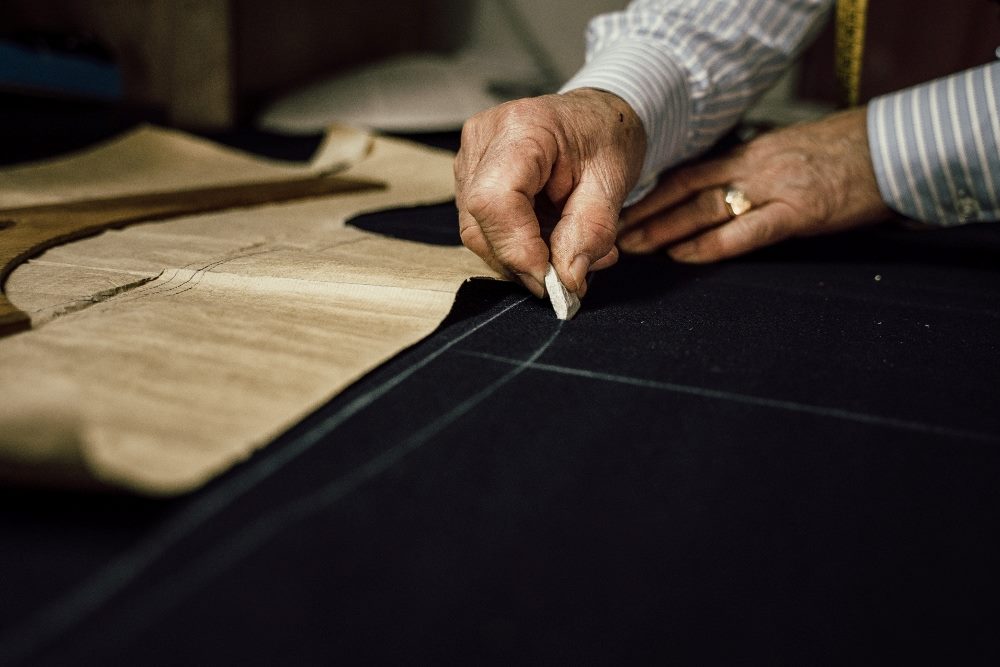 The image depicts an artisan's hands meticulously marking out precise measurements on a dark fabric with chalk. This process symbolizes the precision and personalization akin to "AI personalization" in technology, where algorithms are tailored to meet individual preferences and needs in digital experiences, much like the tailor customizes clothing to fit a specific client perfectly.