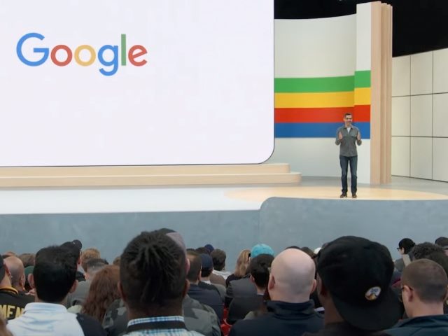 Sundar Pichai on stage at Google I/O 2024 keynote, with "Google" displayed on the large screen to his right, and attendees in the audience in the foreground watching.