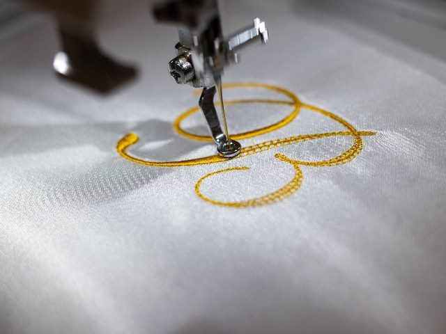 An embroidery machine applies a "B" monogram on a white silk robe with gold thread, suggesting the importance of personalized customer experience. 