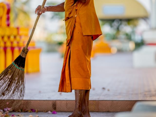 A monk in a saffron robe sweeps temple grounds of garbage with broom.