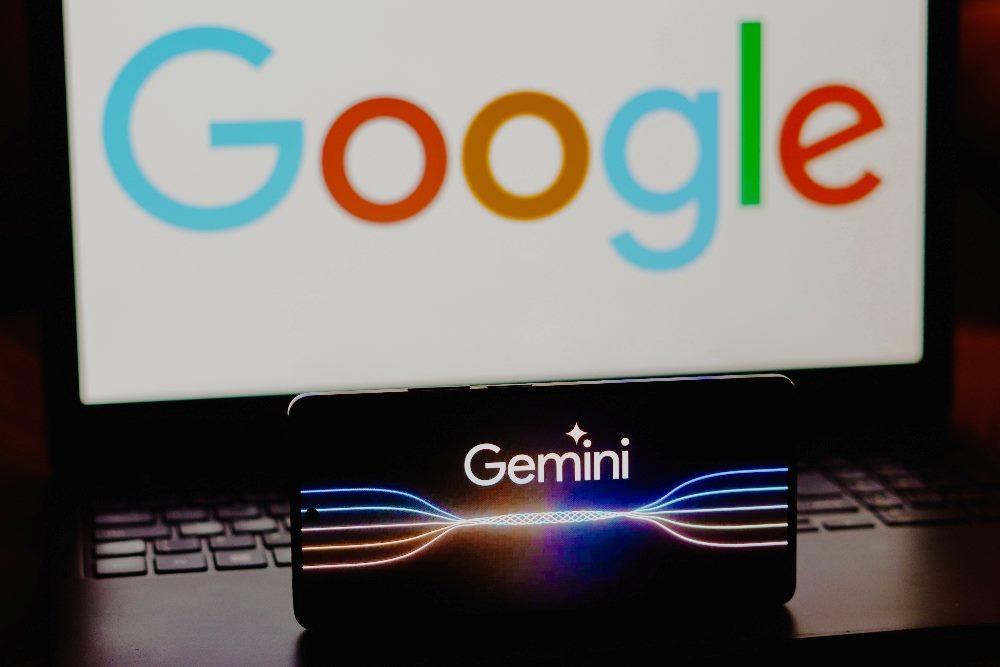 the Google Gemini logo is displayed on a smartphone screen with the Google name in primary colors in the background on a computer screen in piece about Google renaming Bard Google.