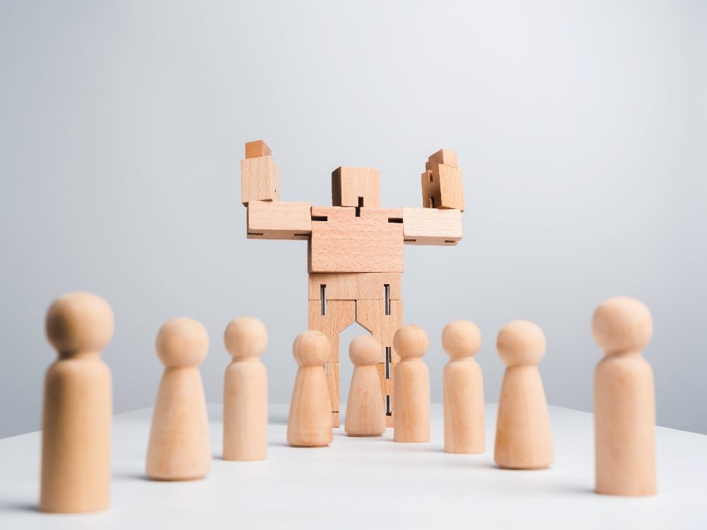 A wooden robot lifts its arms to show its muscles while standing among wooden human follower figures on white background in piece about AI in affiliate and influencer marketing.
