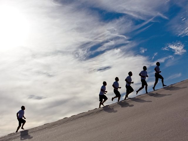 Six men running up a hill, with one man trailing significantly behind.  