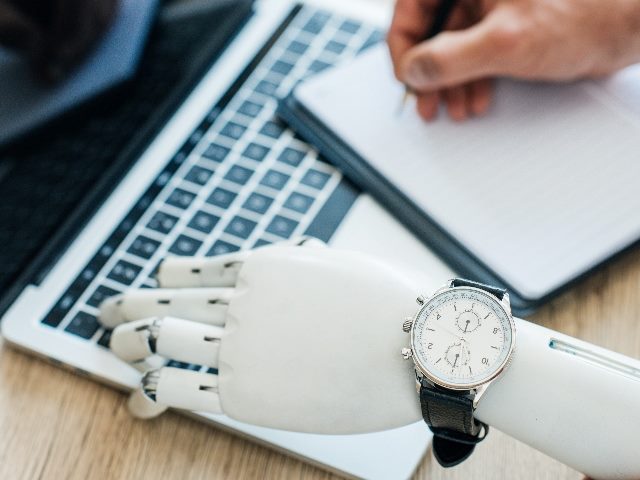 Selective focus of robotic arm with wristwatch using laptop and human hand taking notes at wooden table.