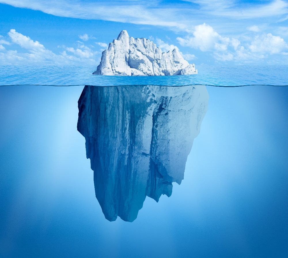 The image shows an iceberg viewed from a side angle under a clear blue sky. The top portion of the iceberg is above water, showing its snowy and jagged surface. Below the waterline, the iceberg extends into a massive, submerged structure, which is larger than the part above water, illustrating the common saying "tip of the iceberg." The entire scene is set against a serene backdrop of sky and calm sea, emphasizing the vastness and hidden depth of the iceberg in piece about Apple reveals at WWDC 2024 and AI development.