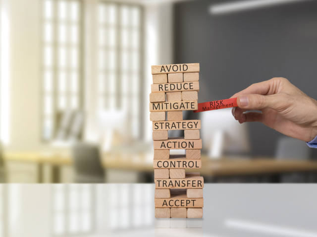 Jenga block game with a hand pulling the risk management block out. There are other words on the other blocks, avoid, control, transfer, etc.