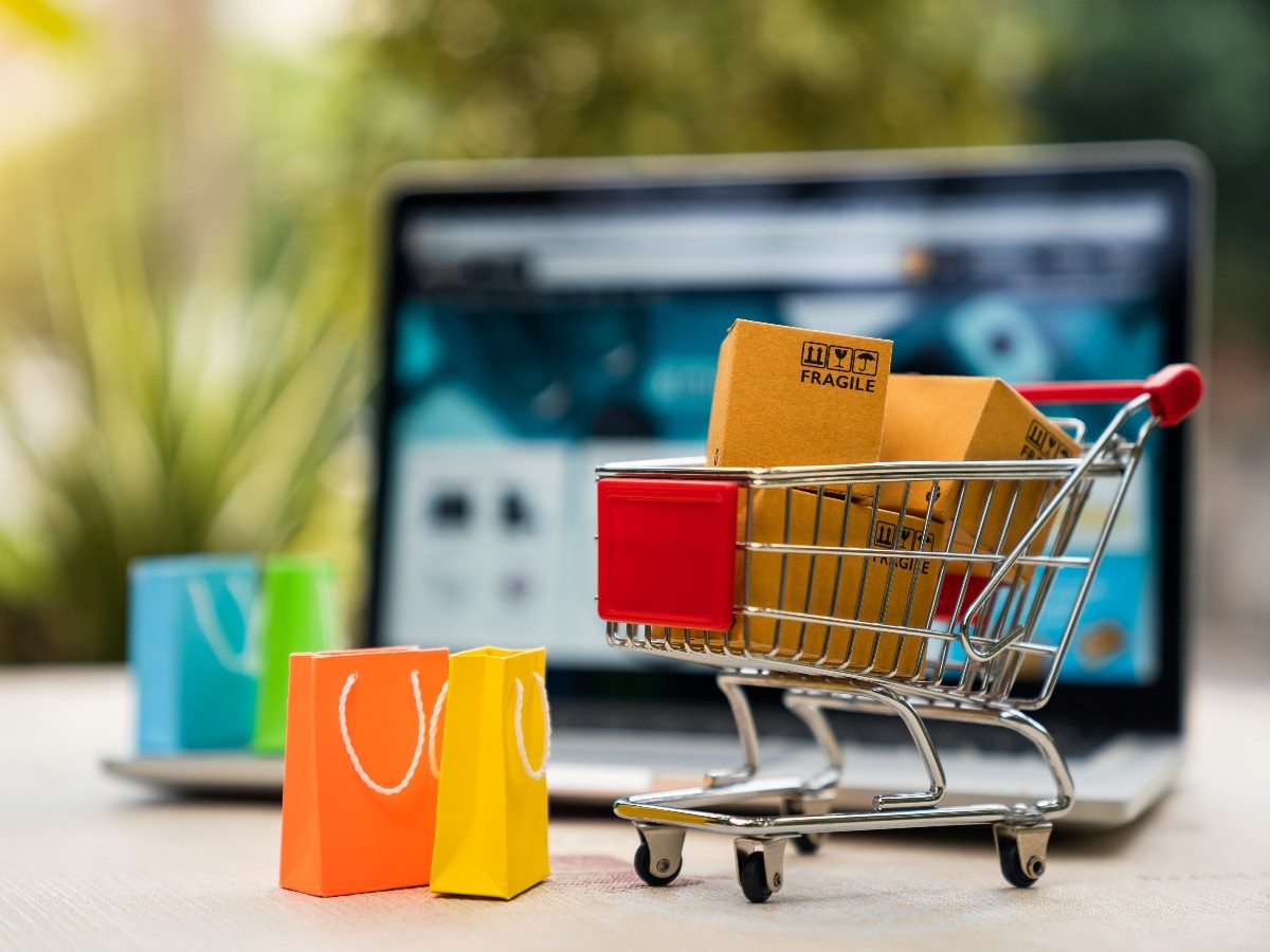 Mini shopping cart holding small cardboard boxes sits in front of an open laptop displaying an ecommerce website, with colorful shopping bags nearby.