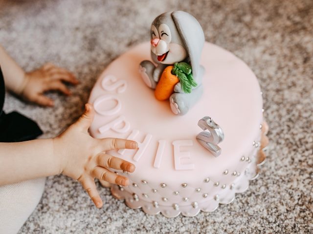 Pink birthday cake name Sophie on it and a rabbit on top and young girl's hands at the side