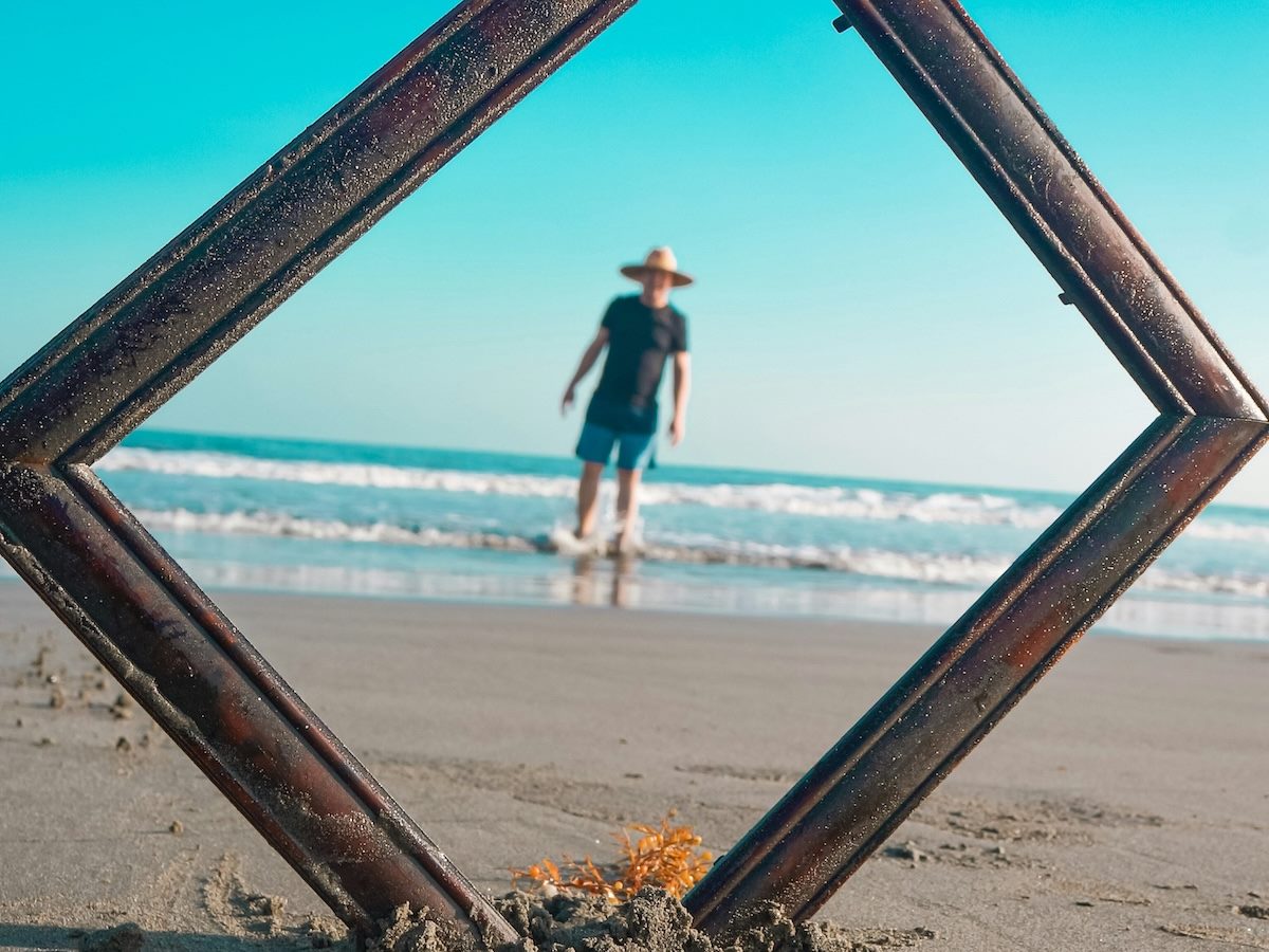 a picture frame stuck in the sand at the beach, a man with a hat on in the center of the frame, ankle deep in the ocean
