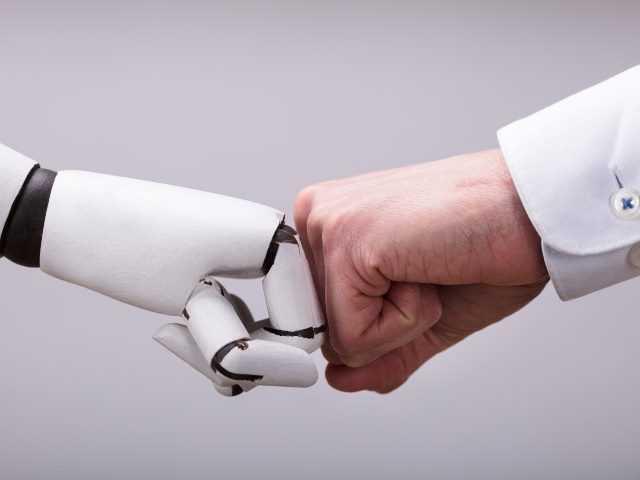 Robot and male human hand doing a fist bump. The man's writs has a white cuff visible. 