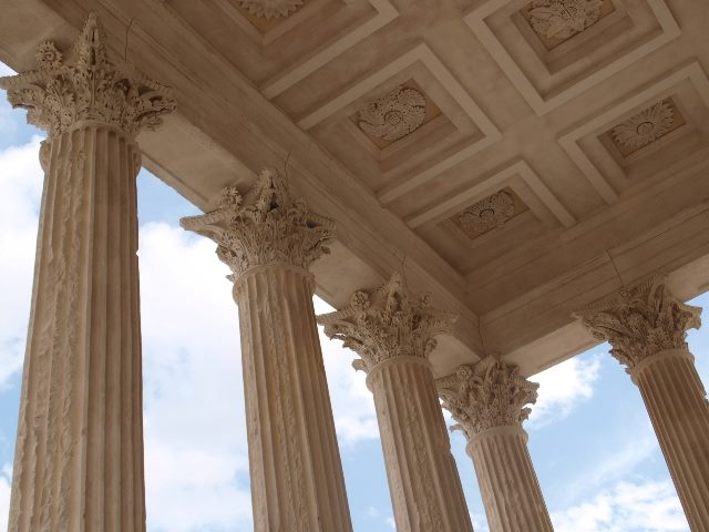 Angled shot of five pillars holding up ornately carved and decorated ceiling of ancient Roman  temple, Maison carrée, in Nimes, France. Each pillar's capital is ornately decorated with flora, leaves and flowers, etc. 
