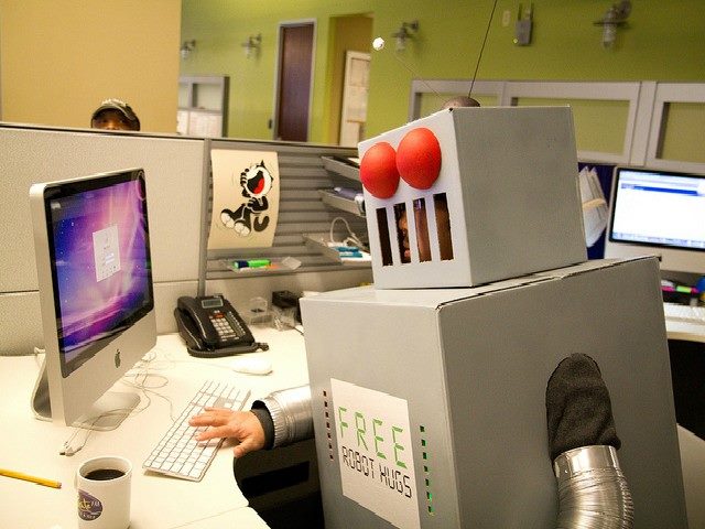 Person dressed up in a robot costume typing on a computer in a cubicle.