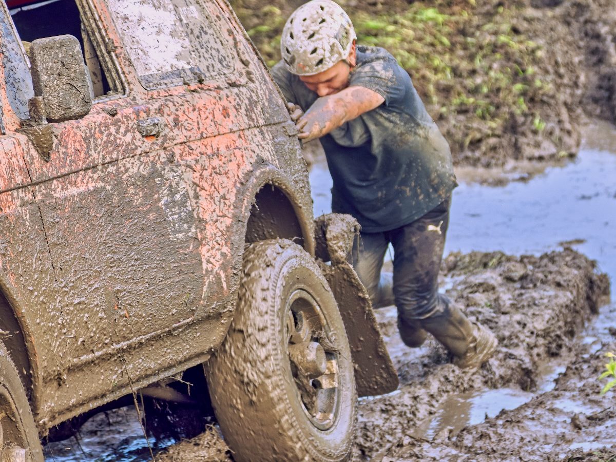 A muddy off-road vehicle stuck in deep mud as a person wearing a helmet and muddy clothes pushes against the front wheel, struggling to free it from a waterlogged trail.