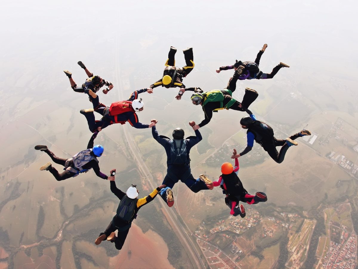 A group of nine skydivers forms a circle while freefalling high above a rural landscape, each person extending arms toward the center in a coordinated formation against a hazy sky.