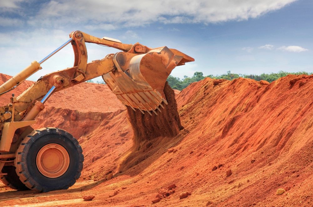 The image depicts an industrial scene of a large yellow mechanical excavator at work in a red soil quarry mining for bauxite for Big Tech products. The excavator's bucket is scooping up a substantial amount of the vividly colored earth, illustrating an active mining operation under a blue sky with sparse clouds. The background includes lush greenery, contrasting sharply with the stark, disturbed red soil foreground. This scene visually captures the impact of industrial activities on natural landscapes in piece about the environmental impact of AI.