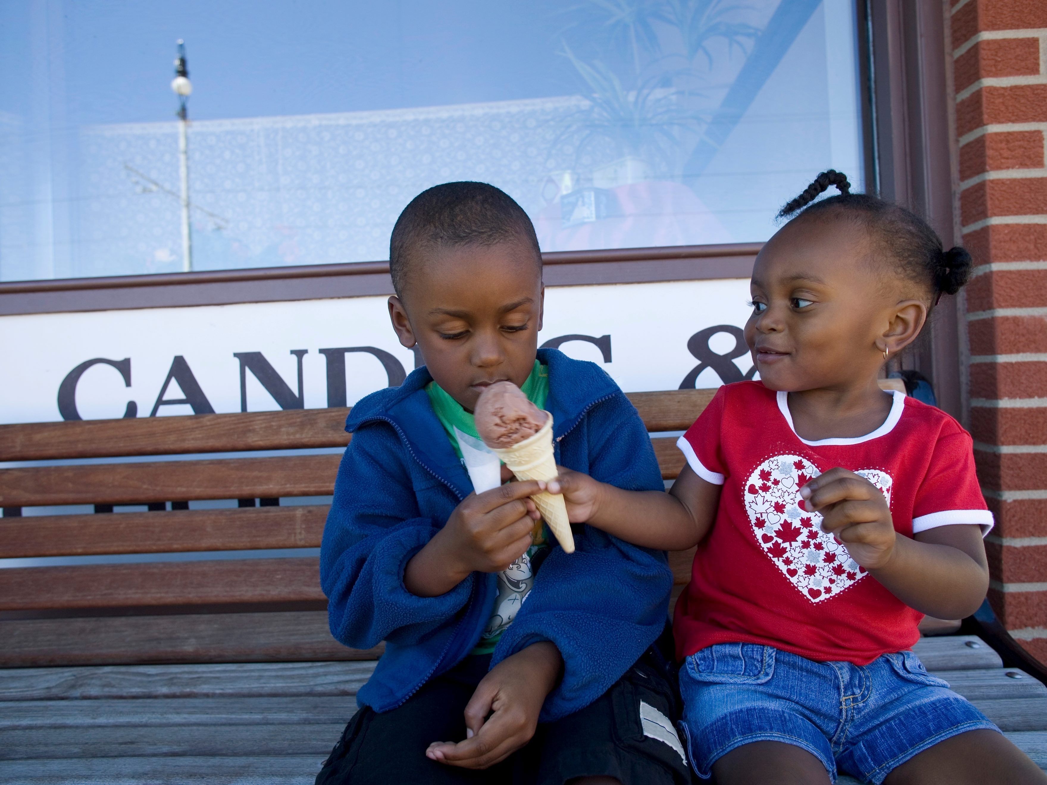 Two children, a boy a girl, sit on a bench with girl in red shirt and braids handing ice cream cone to boy in blue jacket.