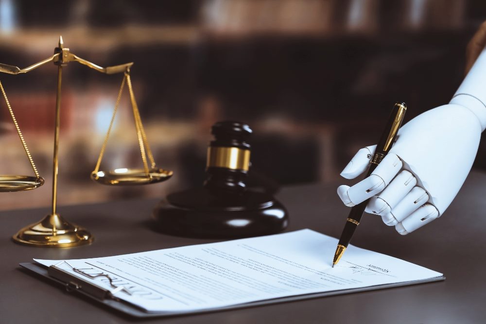 A robot hand holds a pen and signs a legal document on a table next to the scales of justice in piece concerning ethical AI.