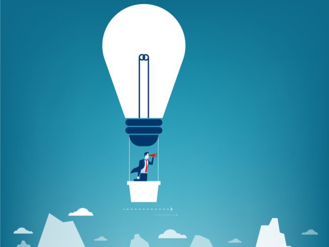 Businessman flying in across blue sky in hot air balloon shaped like a light bulb. Looking over mountain peaks with telescope.