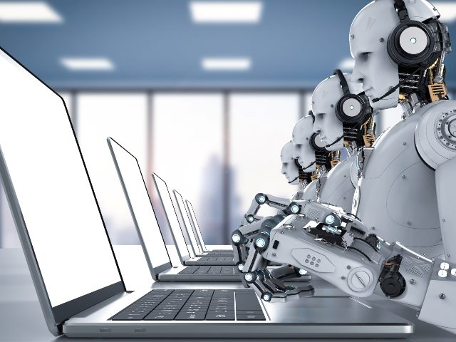 A row of four gray robots wearing headsets work on laptops on a long table. 