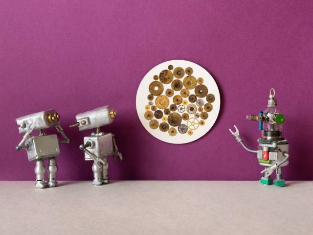 Three toy robots looking at a piece of wall art featuring gears and circles of various brown shades 