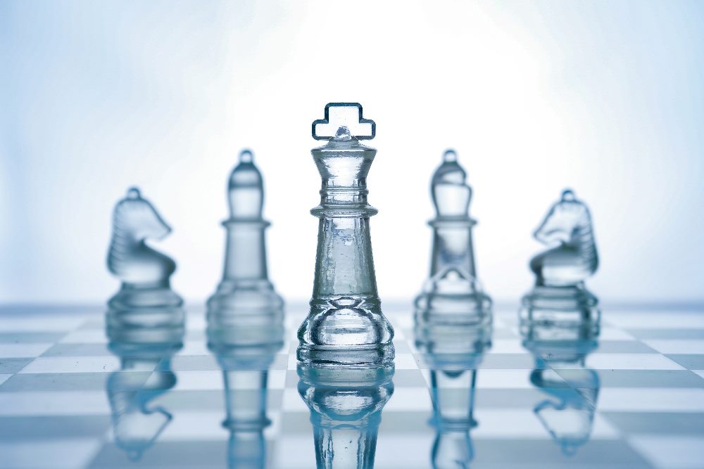 The image shows a set of glass chess pieces arranged on a chessboard, reflecting on the board's surface. In the center stands a tall king, flanked by a queen, bishops, knights, and rooks, all made of clear glass. The pieces are illuminated, casting soft reflections and refractions of light on the glossy surface, highlighting the theme of transparency and strategy. The background is a soft, light gradient, emphasizing the clarity and simplicity of the composition in piece about artificial general intelligence and AI inference.