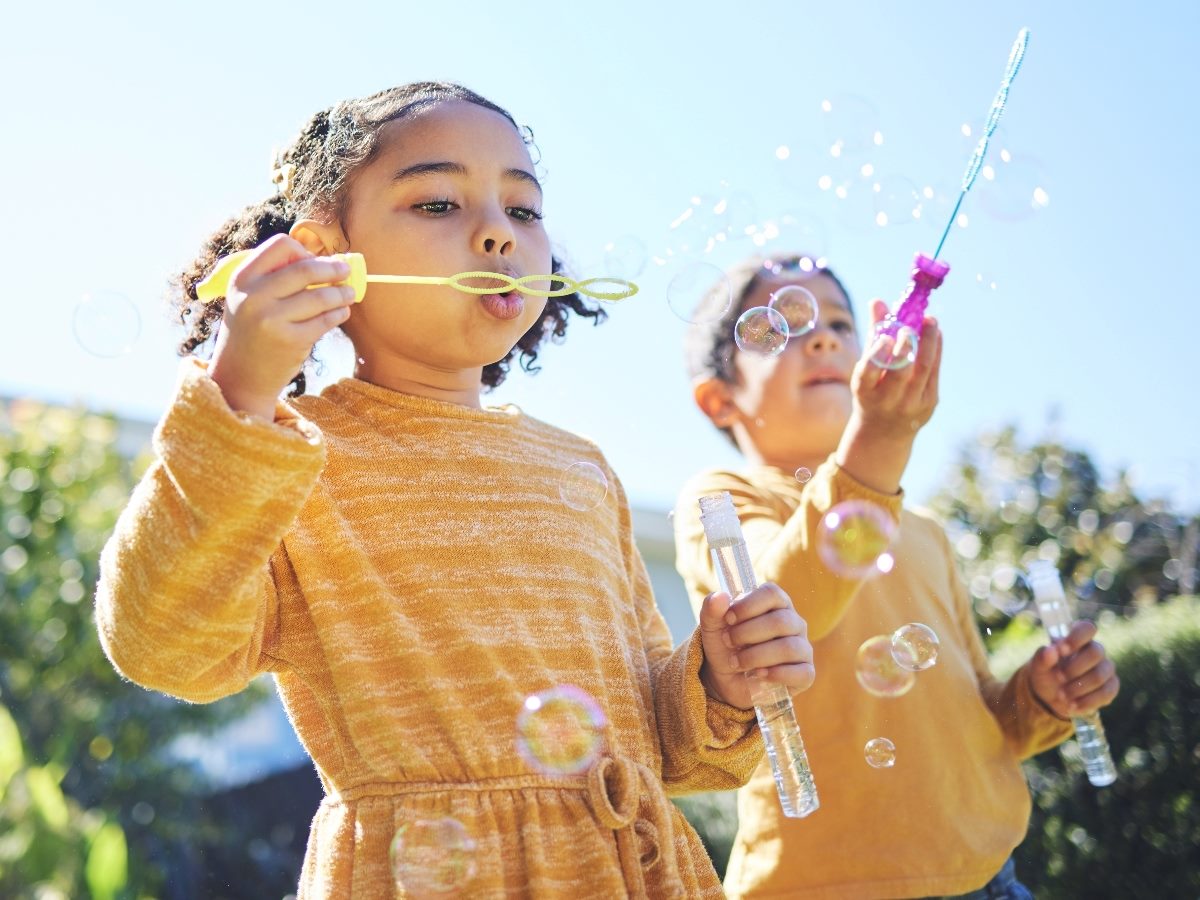Two children wearing yellow sweaters blowing bubbles outdoors on a sunny day, holding bubble wands and bottles.