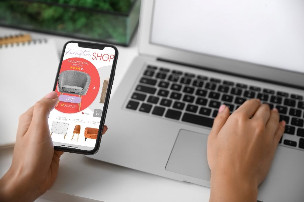 Person with mobile phone and laptop buying online new furniture in piece about the impact of AI integration.