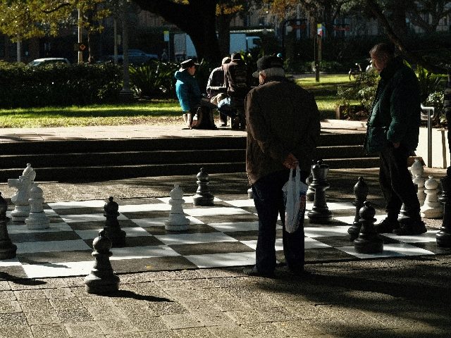 Two men stand on a life-sized chess board.