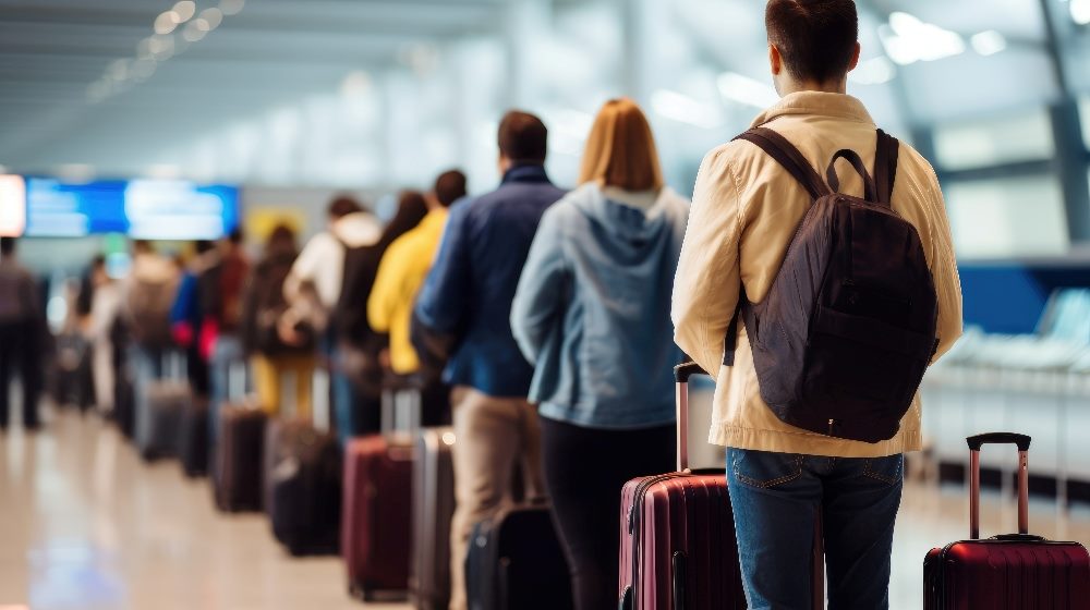 People stand in a a long line with wearing backpacks and carrying their luggage at an airport in piece about AI integration.