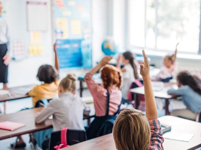 Classroom of children sitting at their desks facing away from the camera hold hands up, with teacher at the front of the classroom and blurred into the background. A globe is also visible in the blurred background.