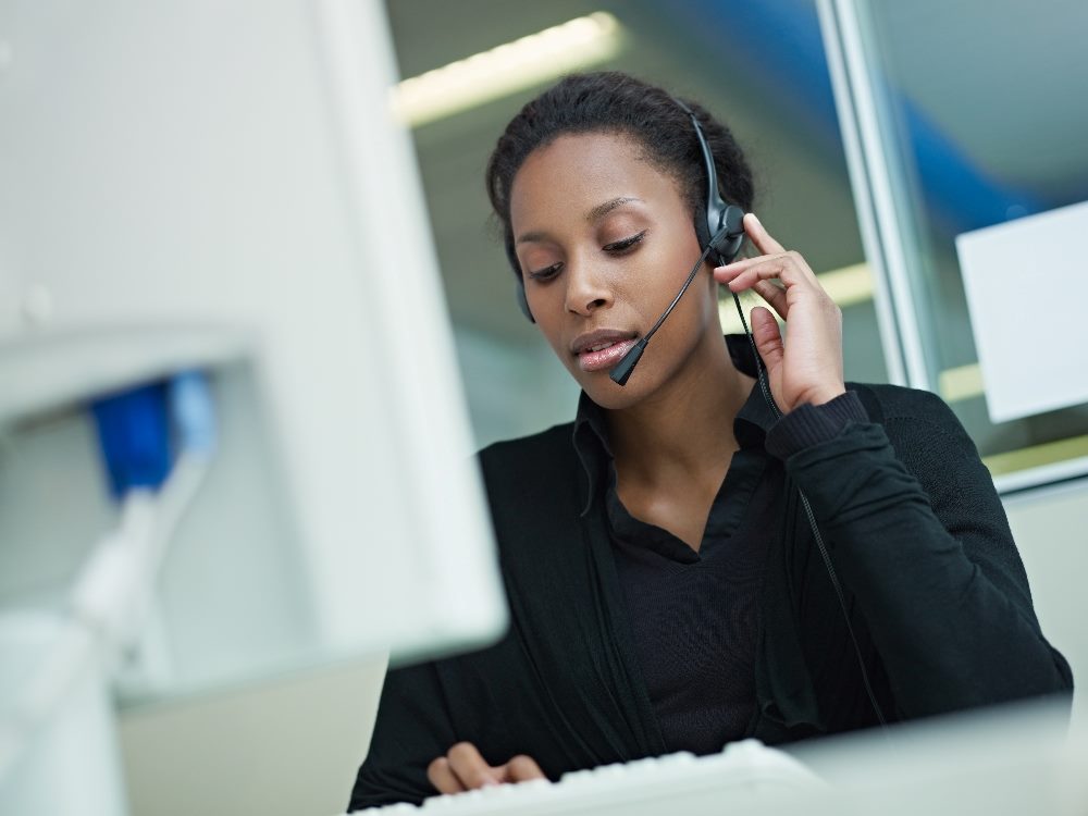 A woman wearing a headset is seated at a desk in a call center. She appears focused, engaged in a conversation, and is typing on a keyboard. The background features office lighting and a computer monitor, indicating a professional work environment in piece about AI in customer service.
