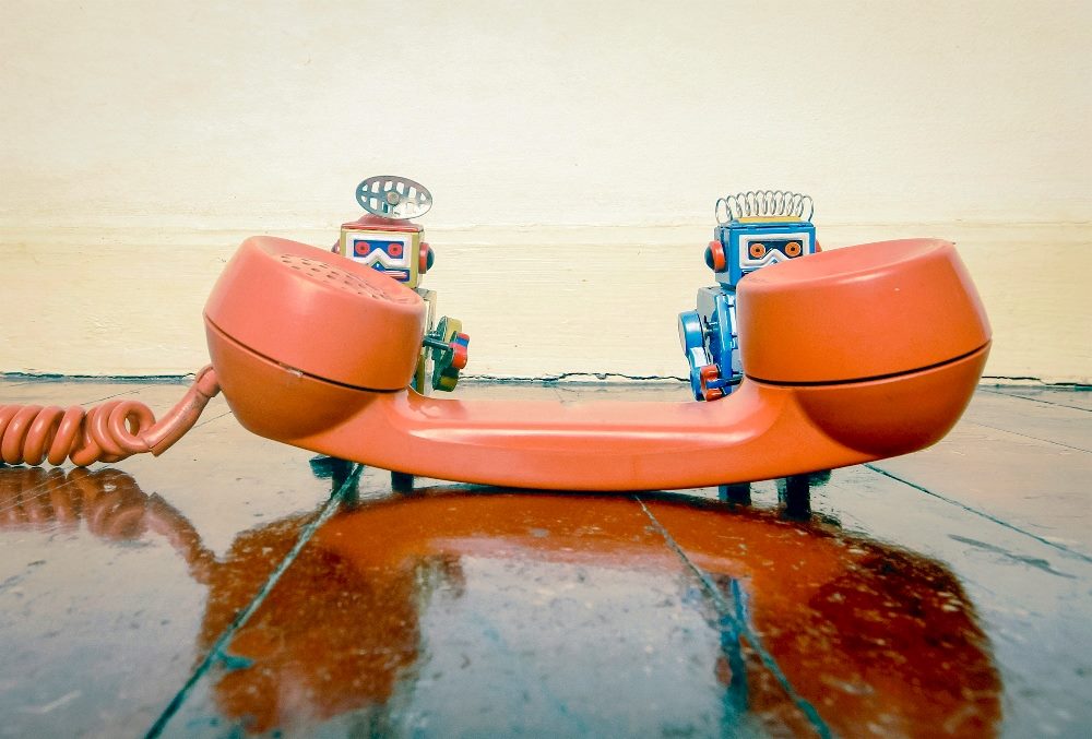 An orange vintage telephone receiver and cord sits on a surface with the receiver facing up and two cute robots, one by the headpiece and one by the mouthpiece, stand behind in in a piece about AI in contact centers.