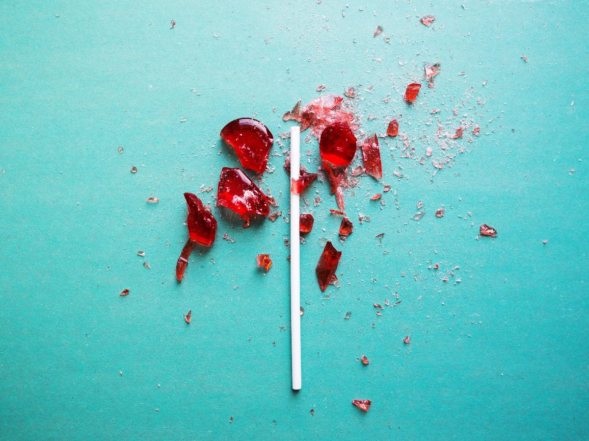 Broken red lollipop shattered into multiple pieces on a teal surface, with fragments and dust scattered around the stick, symbolizing something once whole now disrupted or damaged.