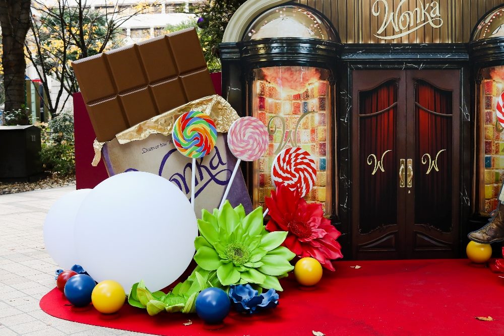 Detail of a Wonka-themed Christmas display with colorful candy's and giant flowers surrounding doors with the name "Wonka" above the doors in Tokyo's Nihonbashi Muromachi area in piece about AI implementation.