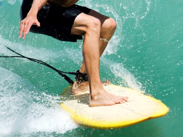 Surfers, torso, legs and one arm and hand visible as he surfs on a yellow surfboard with ankle leash and trying to balance with wave behind him and water splashing around board.
