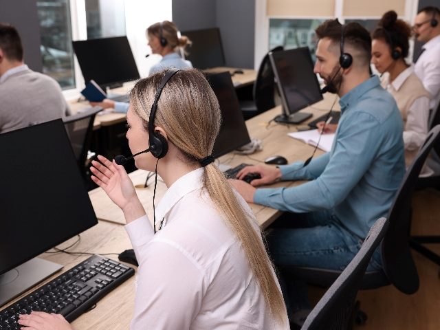 Call center operators working in a modern office seated at long tables with computer screens and keyboards in front of them and wearing headsets, suggesting the improvements of AI in the call center and AI in customer service. 