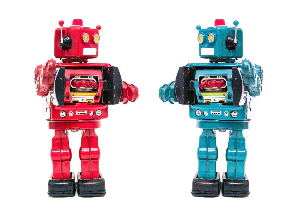 Two vintage-style toy robots, one red and one teal, standing side by side against a plain white background, facing forward with mechanical details and antennae visible.
