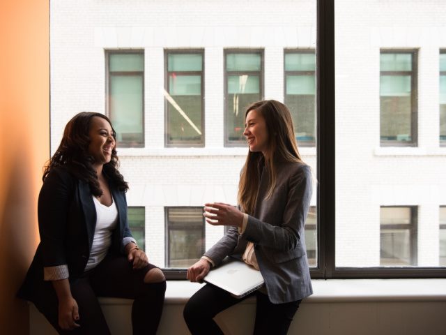 two women sitting together in front of a window having a conversation